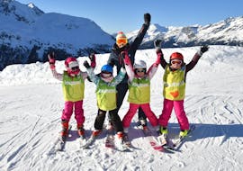 A group of skiers is standing at the top of the slope with their arms in the air with their ski instructor from the ski school ESI Valfréjus during their Kids Ski Lessons (5-12 years) - Holiday - All Levels.